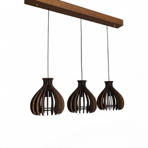May include: Three pendant lights with a dark brown wooden shade in a teardrop shape, suspended from a rectangular wooden beam. The shades have a slatted design, and the cords are black. The background is white.