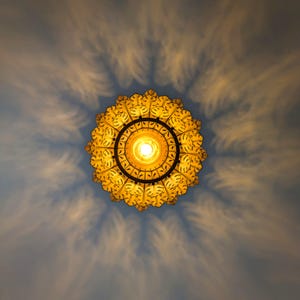 May include: A decorative, ornate ceiling light fixture with a circular design. The light is yellow and casts a pattern of shadows on the blue ceiling. The fixture has a detailed, floral-like pattern around the central light source.