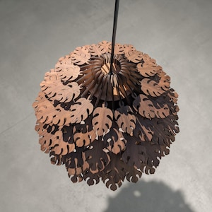 May include: A hanging wooden pendant light fixture. The light is made of layered, laser-cut wooden leaves in a warm brown tone. The leaves are arranged in a spherical shape, with a black cord suspending the light from above. The light casts interesting shadows.