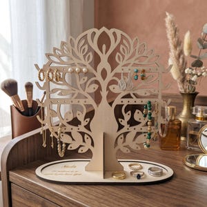 Peut inclure: Un présentoir à bijoux en bois en forme d'arbre, exposant boucles d'oreilles, bracelets et bagues. L'arbre présente un design découpé détaillé avec des feuilles et des branches. La base comporte du texte gravé. Le présentoir est sur une surface en bois.