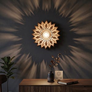 Op de afbeelding: Een houten, zonnebloemvormige wandlamp. Het licht werpt een radiaal patroon op de donkergrijze muur. De armatuur heeft een centrale lichtbron omgeven door gelaagde houten bloemblaadjes. Een houten kast met een plant en decoratie staat eronder.