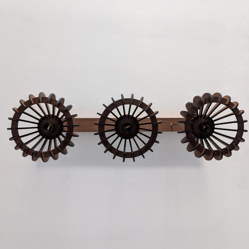 May include: A wooden wall-mounted light fixture featuring three circular, dark brown, spoked shades. The shades are attached to a horizontal wooden beam. The fixture is designed to be mounted on a wall.