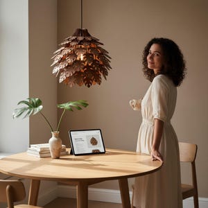 May include: A copper-colored, leaf-shaped pendant light hangs above a round wooden table. A tablet displays a similar light fixture. A woman in a cream-colored dress stands near the table, holding a mug. A vase with a plant is on the table.