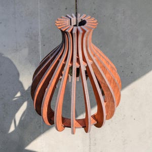 May include: A wooden pendant light fixture with a unique, layered design. The fixture is made of thin, curved wooden slats in a warm brown color, creating an open, organic shape. The light hangs from a thin wire, casting a shadow on the wall.