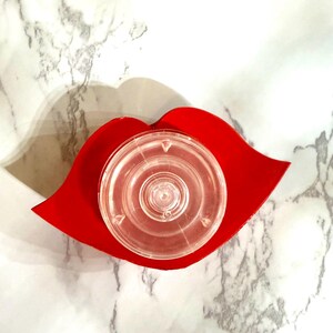 Puede incluir: Un objeto de pl&aacute;stico transparente y circular descansa sobre una bandeja roja con forma de labio. La bandeja es de un rojo vibrante y el objeto de pl&aacute;stico tiene un &aacute;rea central elevada. El fondo es un patr&oacute;n de m&aacute;rmol.