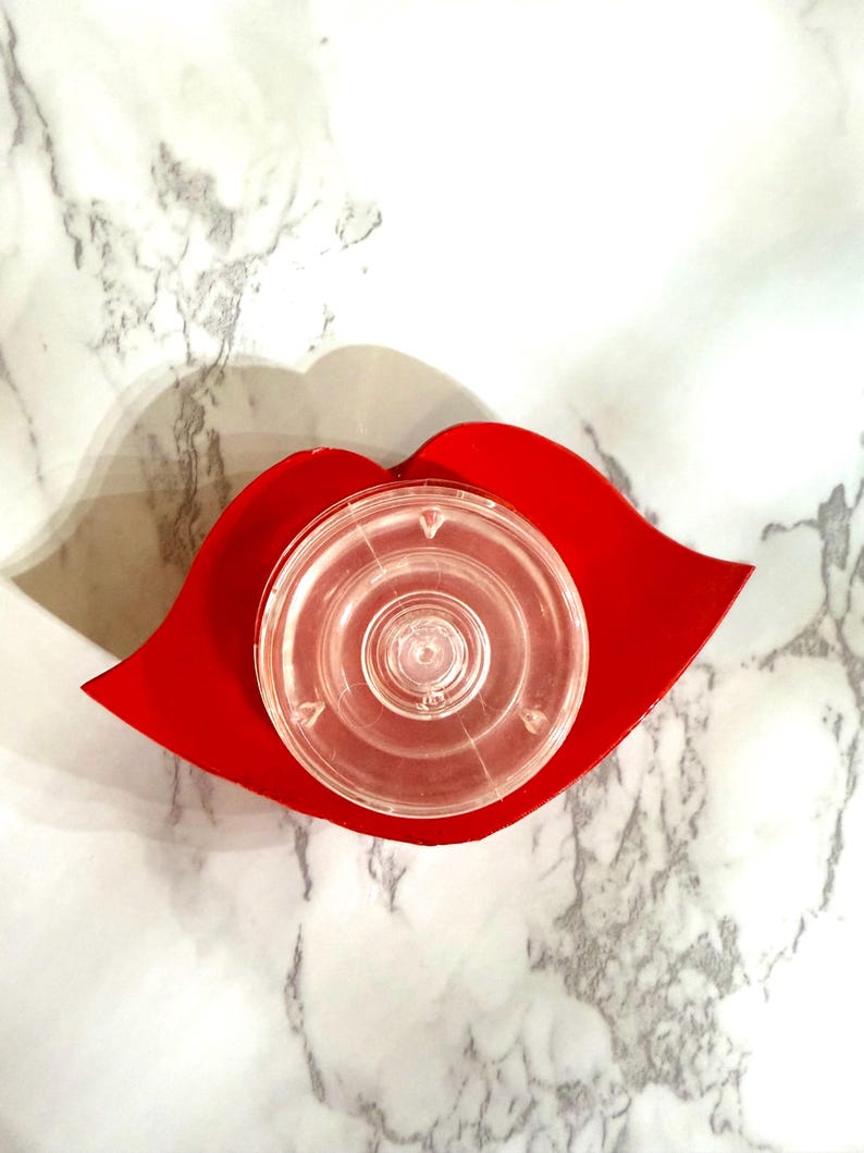 Puede incluir: Un objeto de pl&aacute;stico transparente y circular descansa sobre una bandeja roja con forma de labio. La bandeja es de un rojo vibrante y el objeto de pl&aacute;stico tiene un &aacute;rea central elevada. El fondo es un patr&oacute;n de m&aacute;rmol.