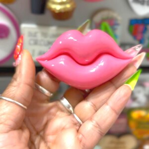 May include: A glossy, pink lip-shaped decorative object. The lips are a vibrant, bubblegum pink color, with a smooth, reflective surface. The object is held in a hand, with other objects in the blurred background.