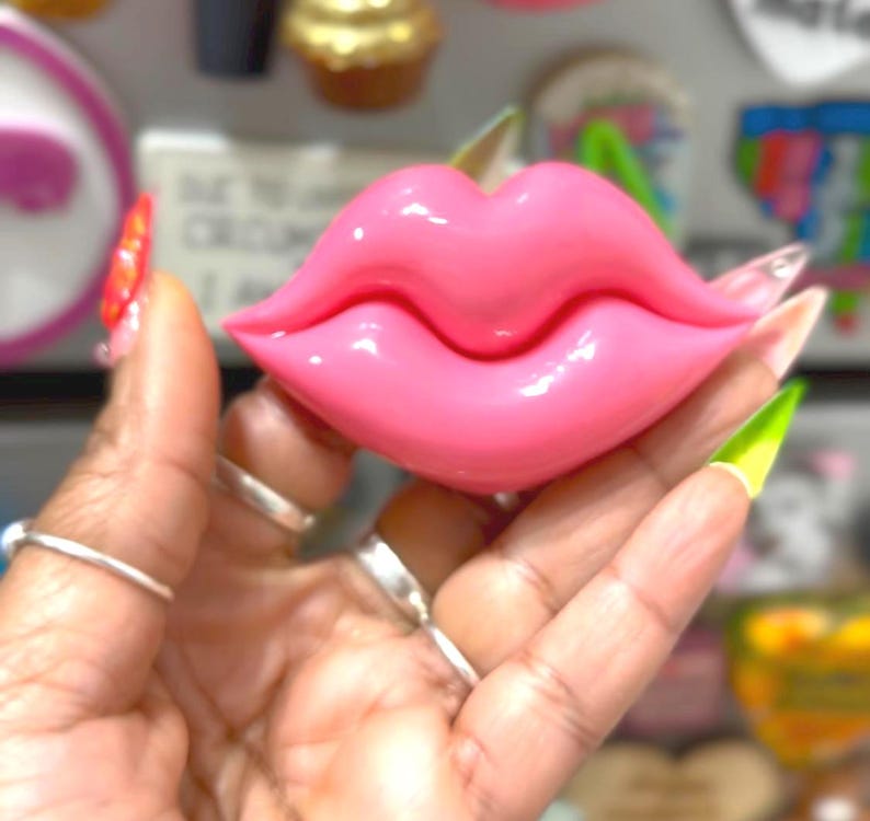 May include: A glossy, pink lip-shaped decorative object. The lips are a vibrant, bubblegum pink color, with a smooth, reflective surface. The object is held in a hand, with other objects in the blurred background.