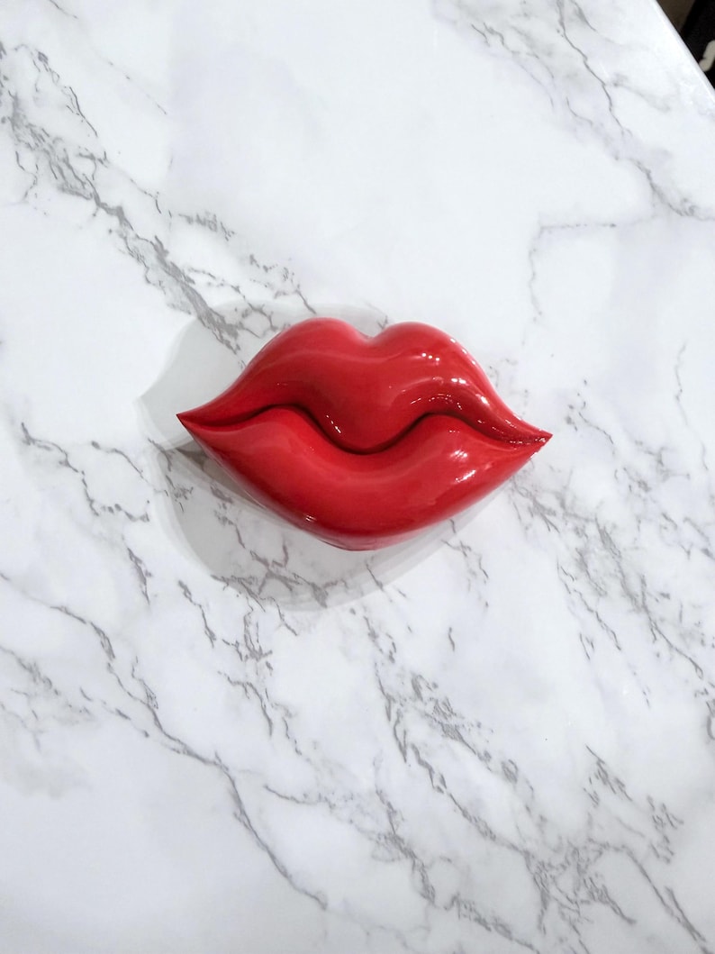 Puede incluir: Un objeto decorativo brillante en forma de labios rojos. Los labios son de un color rojo vibrante y tienen una superficie lisa y reflectante. El objeto est&aacute; colocado sobre una superficie blanca con vetas de m&aacute;rmol.