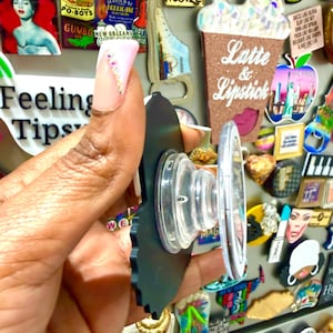 May include: A black phone grip with a clear, circular base is held in front of a collection of colorful magnets. The phone grip has a scalloped edge and a clear, retractable stand. The background includes magnets with text like "Feeling Tips" and "Latte & Lipstick."