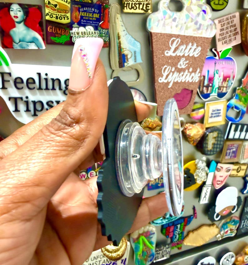 May include: A black phone grip with a clear, circular base is held in front of a collection of colorful magnets. The phone grip has a scalloped edge and a clear, retractable stand. The background includes magnets with text like "Feeling Tips" and "Latte & Lipstick."
