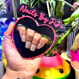May include: A black heart-shaped nail art display with a pink border and the text "Nails By Jifi" in pink. The display is held up to show off glittery nail art. A colorful planter is in the background.