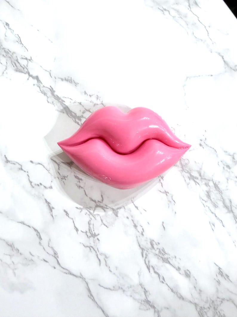 May include: A glossy, pink lip-shaped decorative object. The lips are a vibrant shade of pink and have a smooth, reflective surface. The object is set against a white marble background with gray veining.