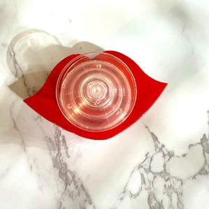 Puede incluir: Un tap&oacute;n de botella de vidrio transparente, circular, con anillos conc&eacute;ntricos, se asienta sobre una base roja en forma de labio. El tap&oacute;n est&aacute; centrado en la base, que es de color rojo vibrante. El fondo es un patr&oacute;n de m&aacute;rmol blanco.