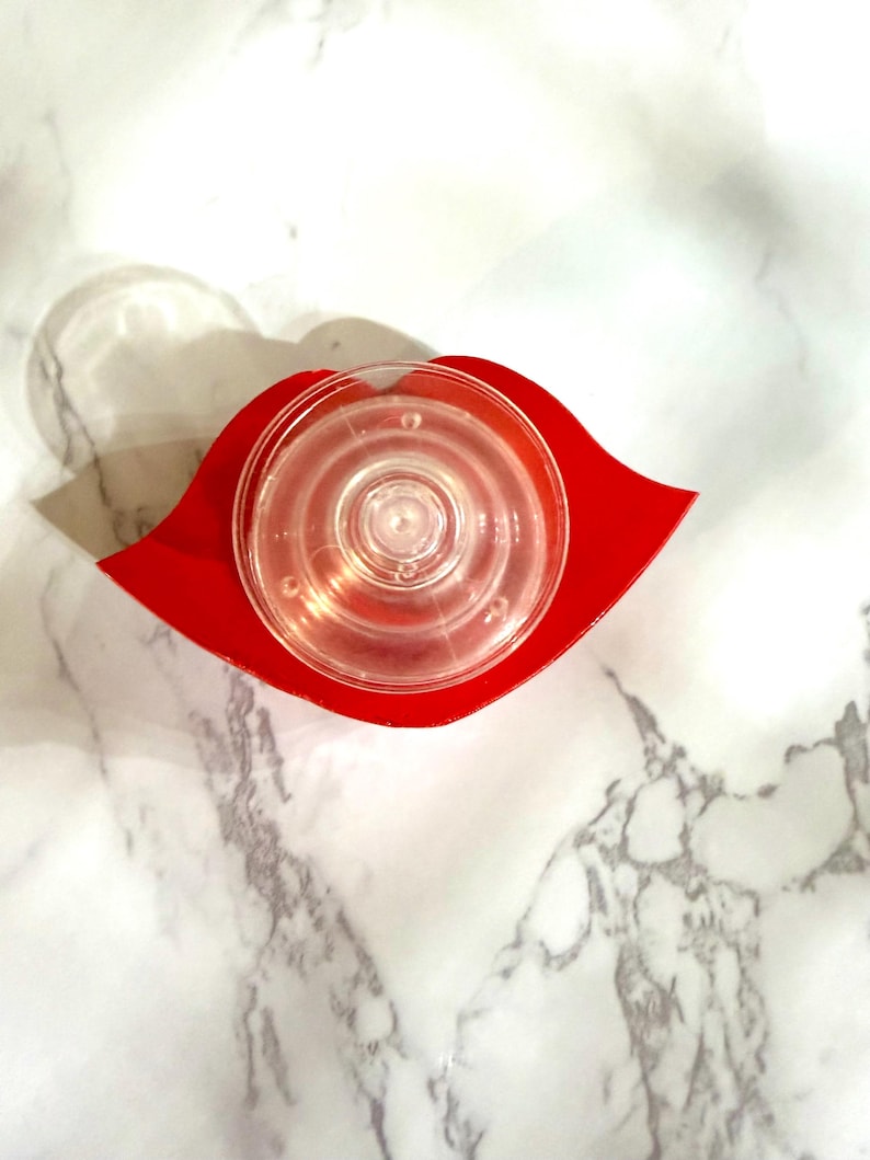 Puede incluir: Un tap&oacute;n de botella de vidrio transparente, circular, con anillos conc&eacute;ntricos, se asienta sobre una base roja en forma de labio. El tap&oacute;n est&aacute; centrado en la base, que es de color rojo vibrante. El fondo es un patr&oacute;n de m&aacute;rmol blanco.