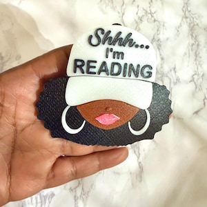May include: A black, brown, and white decorative figure of a person wearing a baseball cap. The cap is white with the words "Shhh... I'm READING" in black. The figure has black hair, brown skin, pink lips, and white hoop earrings.