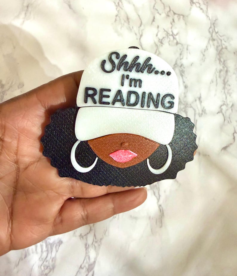 May include: A black, brown, and white decorative figure of a person wearing a baseball cap. The cap is white with the words "Shhh... I'm READING" in black. The figure has black hair, brown skin, pink lips, and white hoop earrings.