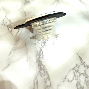 May include: A clear, collapsible phone grip with a black and white top. The grip is designed to attach to the back of a phone or tablet for easier handling and use. The base is clear plastic, and the top has a black border.