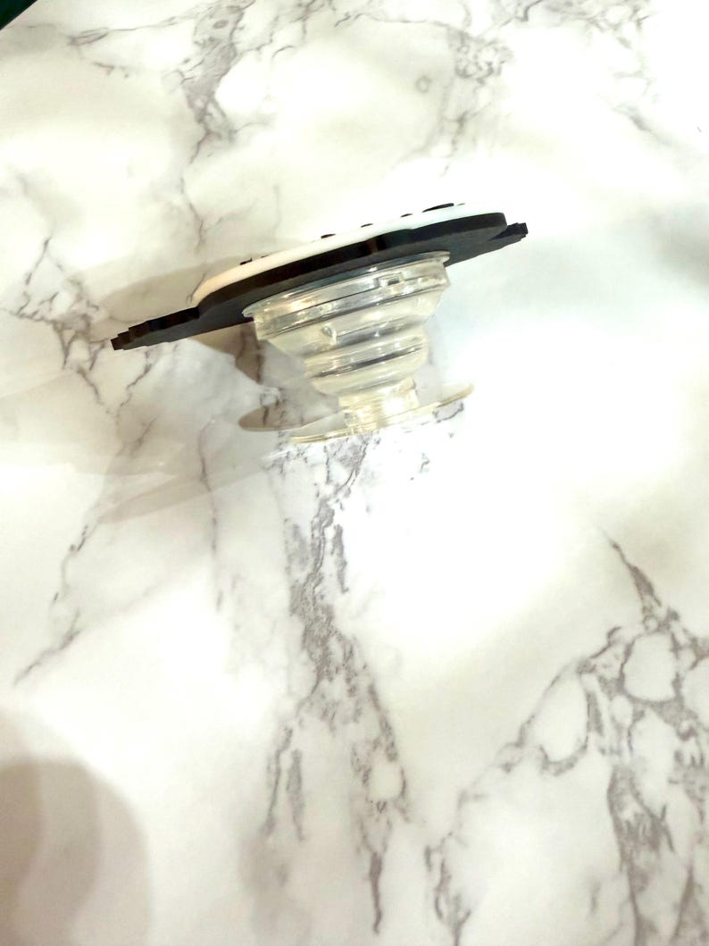May include: A clear, collapsible phone grip with a black and white top. The grip is designed to attach to the back of a phone or tablet for easier handling and use. The base is clear plastic, and the top has a black border.