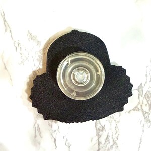 May include: A black, textured, die-cut shape with a clear, circular, plastic object attached. The object has a central button and is designed to be affixed to a surface. The background is a white and gray marble pattern.