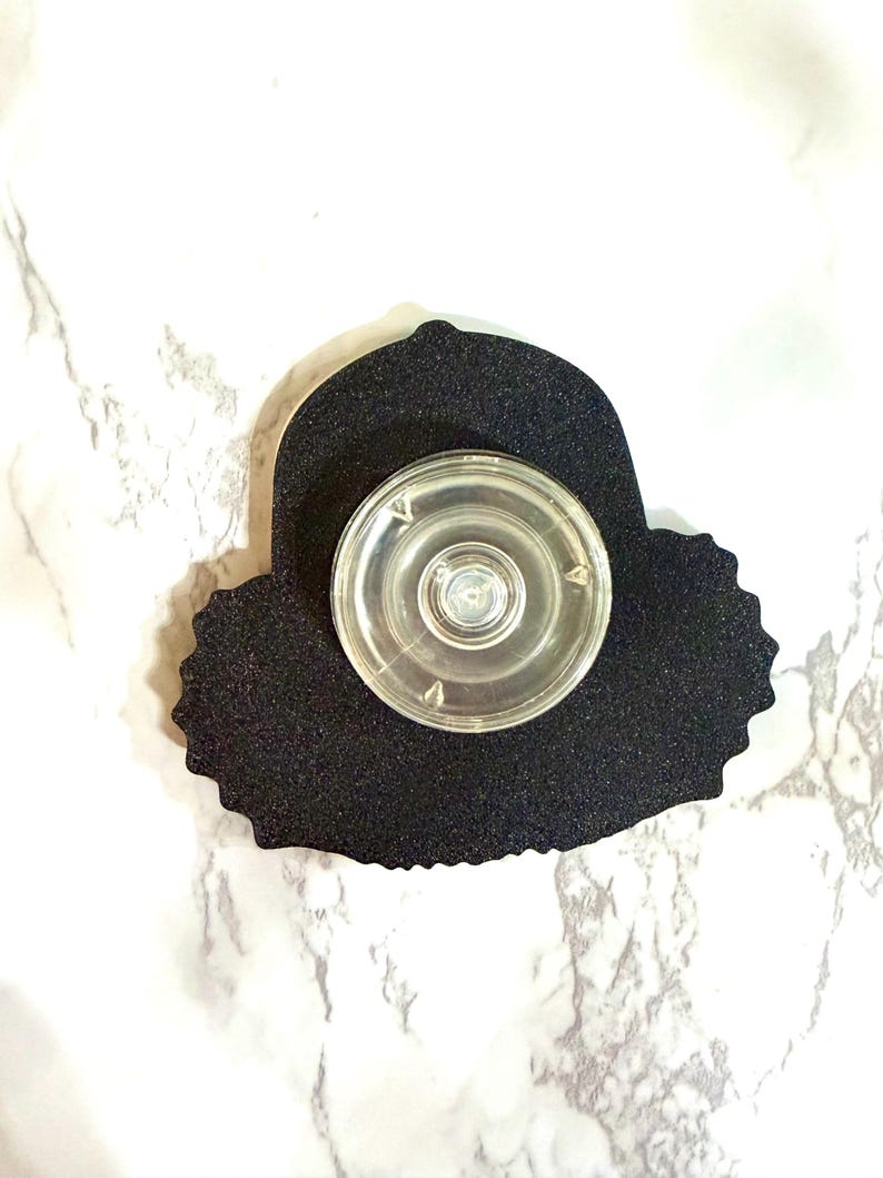 May include: A black, textured, die-cut shape with a clear, circular, plastic object attached. The object has a central button and is designed to be affixed to a surface. The background is a white and gray marble pattern.
