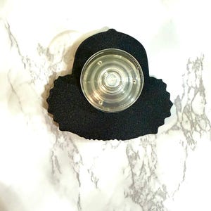 May include: A black phone grip with a clear, circular center. The grip has a textured, irregular edge and is designed to attach to the back of a phone. The background is a white marble pattern.