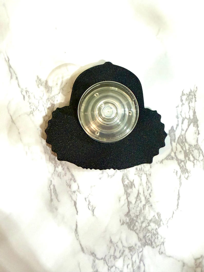 May include: A black phone grip with a clear, circular center. The grip has a textured, irregular edge and is designed to attach to the back of a phone. The background is a white marble pattern.