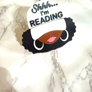 May include: A black and white decorative piece featuring a stylized face with a white cap. The cap has the text "Shhh... I'm READING" in black. The face has brown skin, pink lips, and white hoop earrings. The hair is black.