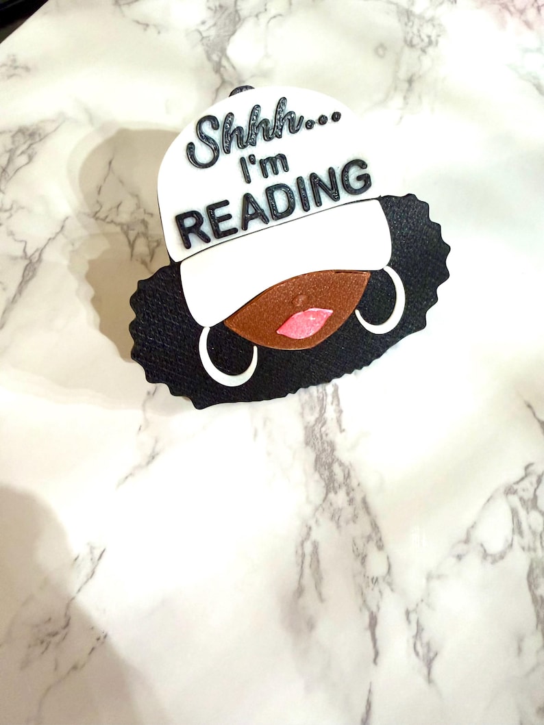 May include: A black and white decorative piece featuring a stylized face with a white cap. The cap has the text "Shhh... I'm READING" in black. The face has brown skin, pink lips, and white hoop earrings. The hair is black.