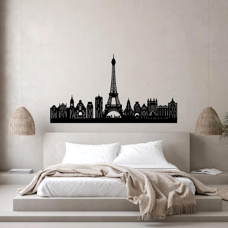 Puede incluir: Arte mural de silueta negra que representa el horizonte de Par&iacute;s, incluyendo la Torre Eiffel y otros edificios ic&oacute;nicos. La obra de arte est&aacute; montada sobre una cama con ropa de cama blanca y una manta de tonos neutros. Luces colgantes decorativas a ambos lados.