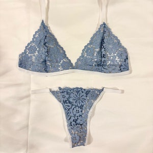 May include: A blue lace lingerie set featuring a triangle bralette and a thong. The bralette has white straps and trim, while the thong has white trim. The lace has a floral pattern.