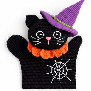 May include: A black crochet cat puppet wearing a purple witch hat with a green band. The cat has white whiskers, eyes, and a pink nose. An orange ruffled collar and a white spiderweb design complete the Halloween-themed puppet.