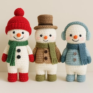 Op de afbeelding: Drie handgemaakte gehaakte sneeuwmannen. De sneeuwmannen zijn wit met zwarte knoopogen en oranje wortelneuzen. Elke sneeuwman draagt een andere outfit: een rode hoed en sjaal, een bruine hoge hoed en jas, en een blauwe oorwarmers en jas. Ze zijn ongeveer 15 cm hoog.