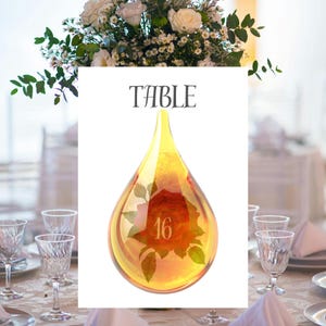 May include: A decorative table number featuring a teardrop-shaped glass design with a red rose and the number 16 inside. The word "TABLE" is printed at the top. The table setting includes glassware and a floral arrangement.