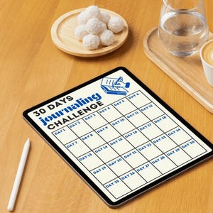 May include: A white tablet with a 30-day journaling challenge grid. The grid is divided into 30 squares, each labeled with a day number. The tablet is on a wooden surface with a glass of water and a plate of white chocolate truffles.