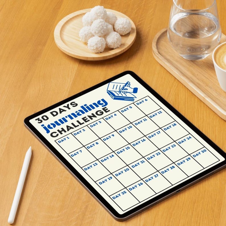 May include: A white tablet with a 30-day journaling challenge grid. The grid is divided into 30 squares, each labeled with a day number. The tablet is on a wooden surface with a glass of water and a plate of white chocolate truffles.