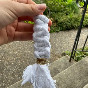 K&ouml;nnte beinhalten: Wei&szlig;er geflochtener Schl&uuml;sselanh&auml;nger aus Seil mit einem goldenen Ring und einem wei&szlig;en Quasten.