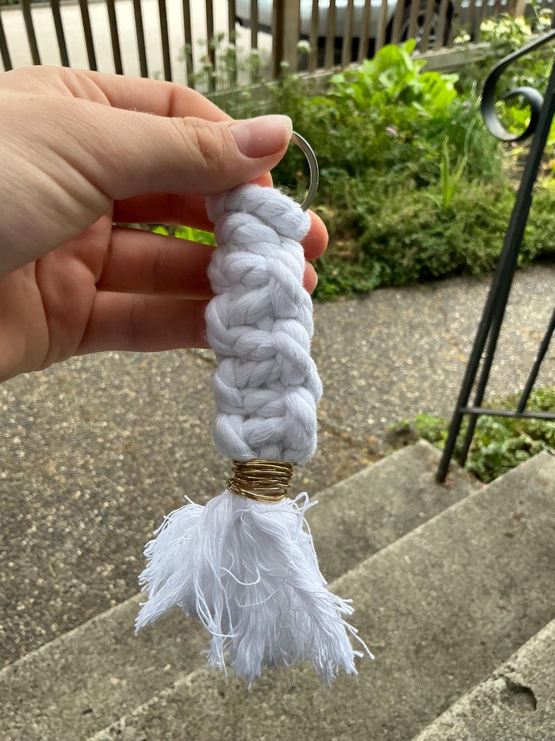 K&ouml;nnte beinhalten: Wei&szlig;er geflochtener Schl&uuml;sselanh&auml;nger aus Seil mit einem goldenen Ring und einem wei&szlig;en Quasten.