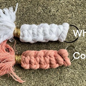 K&ouml;nnte beinhalten: Zwei handgefertigte Makramee-Schl&uuml;sselanh&auml;nger mit Quasten. Einer ist wei&szlig; und der andere korallenfarben.