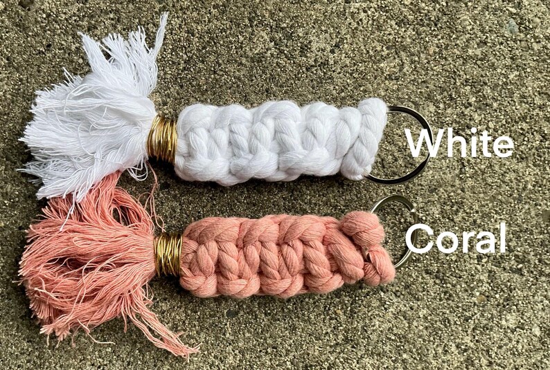 K&ouml;nnte beinhalten: Zwei handgefertigte Makramee-Schl&uuml;sselanh&auml;nger mit Quasten. Einer ist wei&szlig; und der andere korallenfarben.