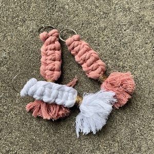 K&ouml;nnte beinhalten: Drei rosafarbene und wei&szlig;e geh&auml;kelte Schl&uuml;sselanh&auml;nger mit Quasten. Die Schl&uuml;sselanh&auml;nger sind aus weichem, flauschigem Garn gefertigt und haben ein schlichtes, elegantes Design.