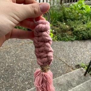 K&ouml;nnte beinhalten: Ein rosa geflochtener Schl&uuml;sselanh&auml;nger mit einem goldenen Metallring und einem rosa Quaste.
