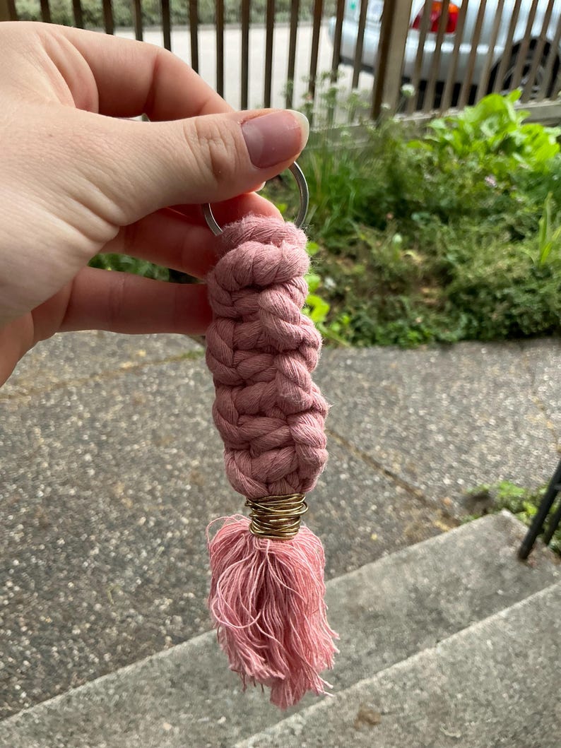 K&ouml;nnte beinhalten: Ein rosa geflochtener Schl&uuml;sselanh&auml;nger mit einem goldenen Metallring und einem rosa Quaste.
