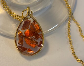 May include: A gold-colored chain necklace with a teardrop pendant. The pendant has a gold frame and is filled with orange-colored, irregularly shaped pieces. The necklace is displayed on a white surface.