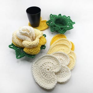 May include: A collection of handmade crochet items, including coasters and a cup holder. The coasters are in various shapes and colors, including yellow, white, and green. A black cup sits on a yellow coaster. The items are designed to protect surfaces.