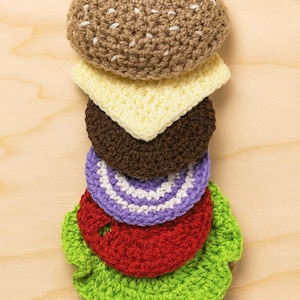 May include: A crocheted burger with a sesame seed bun, cheese, a brown patty, purple onion, red tomato, and green lettuce. The handmade burger is stacked on a light wood surface.