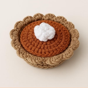 May include: A handmade crochet pumpkin pie with a dollop of white cream. The pie features a brown crust, a pumpkin-colored filling, and a white cream topping. The pie is a cozy, handcrafted item.