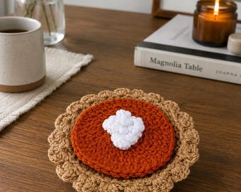 May include: A round, crocheted coaster designed to resemble a pumpkin pie. The coaster features a tan, scalloped crust edge, a dark orange center representing the filling, and a small, textured white cluster in the middle resembling whipped cream.