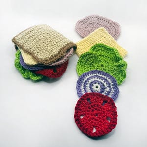May include: A collection of colorful, crocheted food items, including a sandwich and various ingredients. The sandwich features tan bread, with layers of green lettuce, purple, yellow, and red components. Additional crocheted items include a purple heart, yellow square, and green circle.