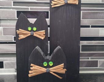 May include: Three black wooden cat figures of varying heights. Each cat has green gem eyes, a raffia bow tie, and a small gray nose. The cats are displayed against a gray and white tiled background.
